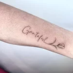 Fine line tattoo reads "Grateful" in cursive with tulip at the end.
